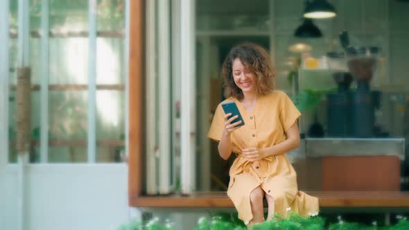 Laughing Curly Woman in Yellow Dress Using Her Phone While Sits in Coffee Shop