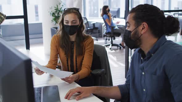 Diverse male and female colleague wearing face masks sitting at desk holding document in discussion