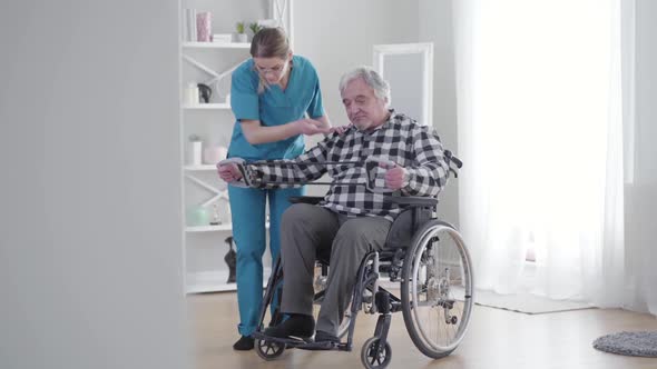 Long Shot of Concentrated Caucasian Woman Helping Old Man To Stretch Elastic Band, Mature Retiree