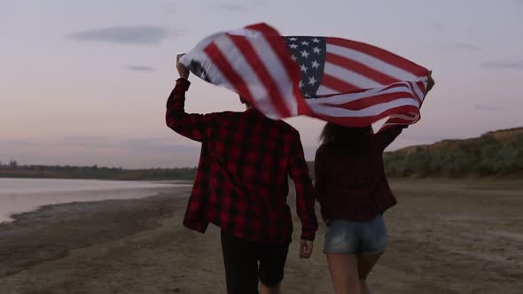 Young Couple Both in Red Plaid Shirts are Running on the Seashore with American Flag Raised Above