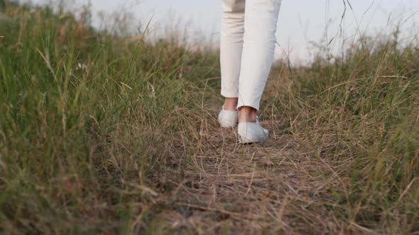 The Legs of a Woman Dressed in White Clothes Walk Along the Path Among the Grass