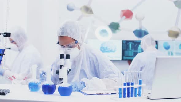 Female Scientist in Coverall Equipment Writing Notest and Looking Through Microscope