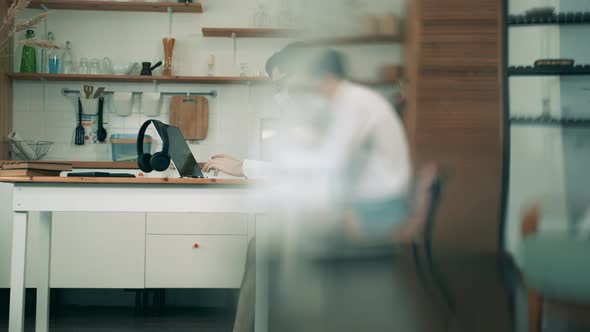 A Man in a Protection Mask Is Working on a Laptop in the Kitchen