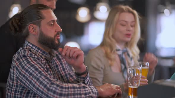 Bearded Smiling Man Talking with Blurred Friends Sitting at Bar Counter in Pub Indoors