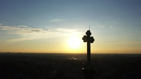 An aerial shot of a cathedral's steeple with a cross on top, taken at sunrise. The camera track left