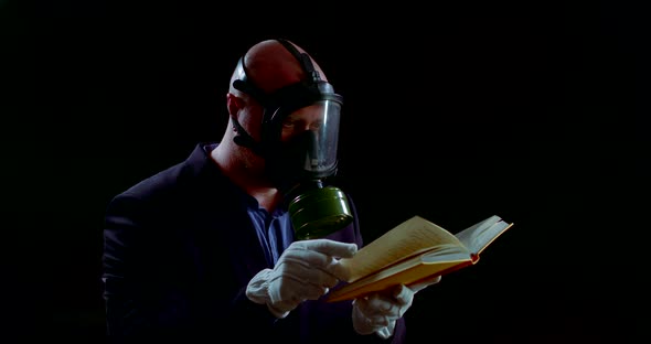 Man with Respirator on Face and Protective Gloves on Hands Is Flipping Page of Book, Reading