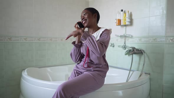 Confident Beautiful African American Woman in Pajamas Sitting on Bathtub Talking on Phone Smiling