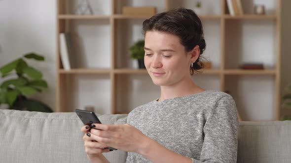 Young Woman Relax on Couch at Cozy Home Using Cellular Looking Pensive Thinks Over Purchase on