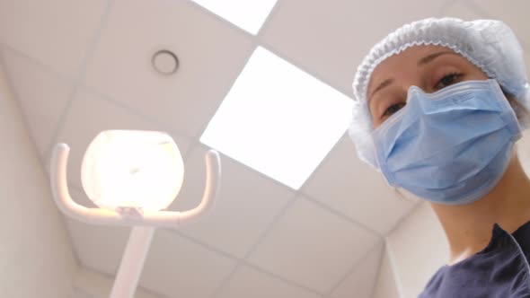 Beautiful Woman Dentist Turns on a Lamp and Stands Over a Patient with a Tongs for Removing Teeth