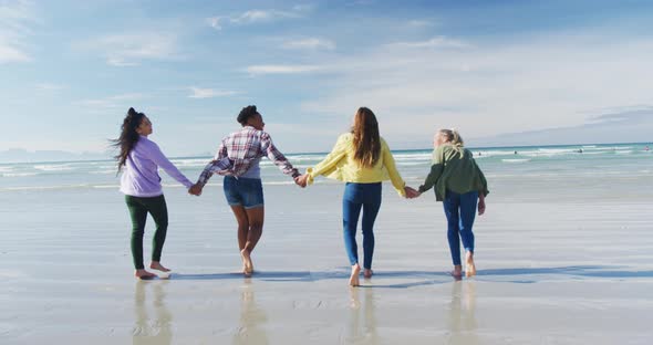 Happy group of diverse female friends having fun, walking along beach holding hands