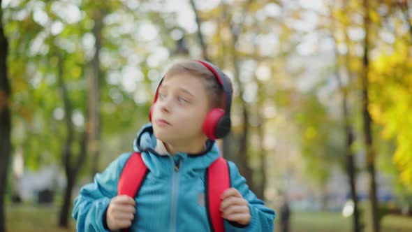 Cheerful Caucasian Schoolboy in Headphones Dancing Walking with Backpack in Sunny Autumn Park