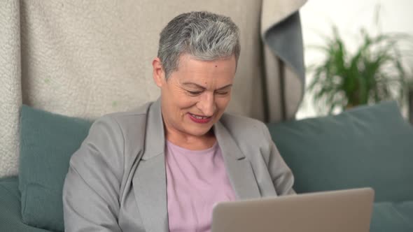 Mature Woman with a Gray Short Haircut is Working Sitting on the Couch with a Laptop