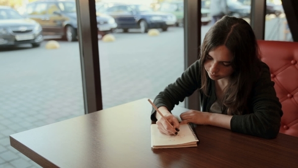 Teenage Student Makes Sketches In Notebook In Cafe