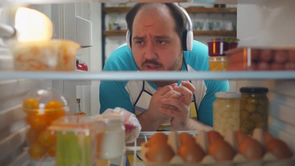 View From Fridge of Cheerful Overweight Man in Apron and Headphones Taking Eggs From Shelf