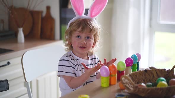 Cute two-year-old boy in bunny ears paint Easter eggs with multi-color paints sitting at the table