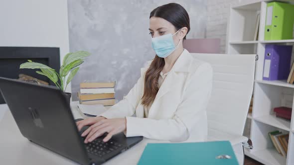 Side View Portrait of Young Concentrated Woman in Covid Face Mask Typing on Laptop Keyboard Sitting