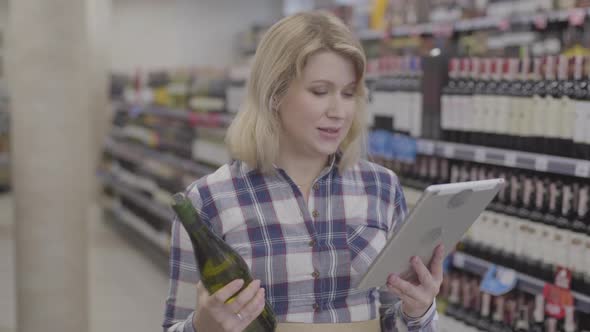 Young Cheerful Caucasian Woman Comparing Information in Tablet with Label on Wine Bottle. Positive