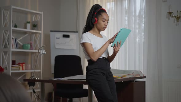 Portrait of Serious Young African American Woman in Headphones Sitting on Table Surfing Internet on