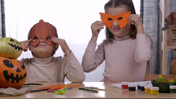 A Brother and Sister are Sitting at the Table in the Room Trying on Face Masks Cut Out of Paper in