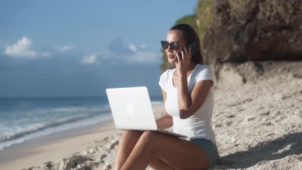 Girl Freelancer in Sunglasses Works on Beach with Laptop and Talks on Phone