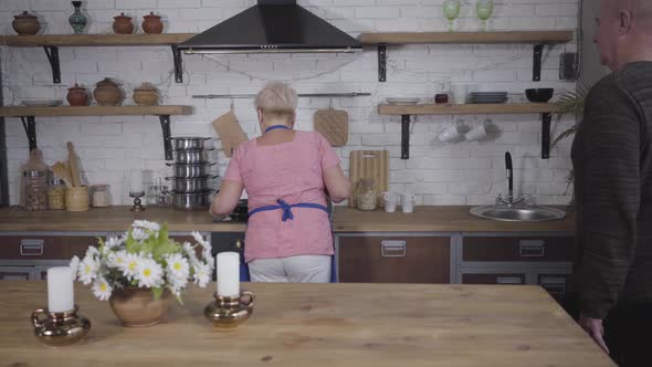 Back View of Mature Bald Caucasian Senior Man Approaching His Wife and Presenting Her Bouquet