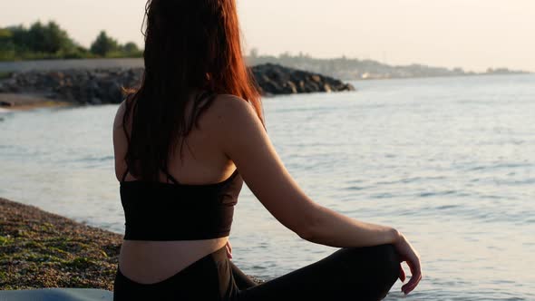 A yogi woman sits in a lotus position on the seashore and looks at the sea dawn of the sun.