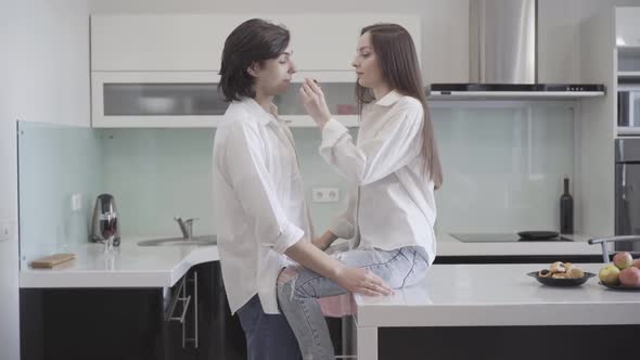 Charming Millennial Woman Sitting on Tabletop in Kitchen Feeding Man with Sweet Delicious Bakery