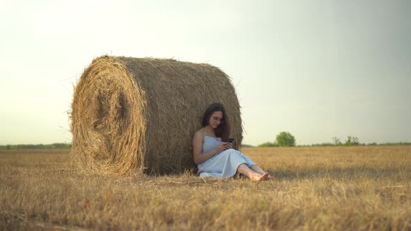 A Young Woman Sits Near a Haystack in a Field with a Phone in Her Hands