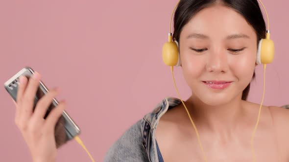 Happy pretty Asian teenager girl dancing and listening music by smartphone on pink background.