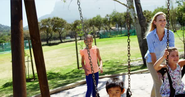 Happy trainer pushing schoolkids on swing in playground