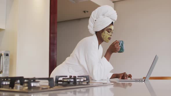 African american attractive woman with applied face mask drinking coffee and using laptop in kitchen