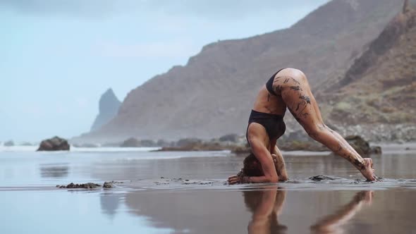 A Beautiful Woman in a Swimsuit Performs a Headstand on a Beach with Black Sand Near the Sea