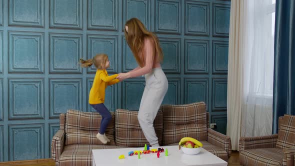 Young Mother Babysiter and Little Kid Daughter Jumping Dancing on Sofa in Modern House Living Room