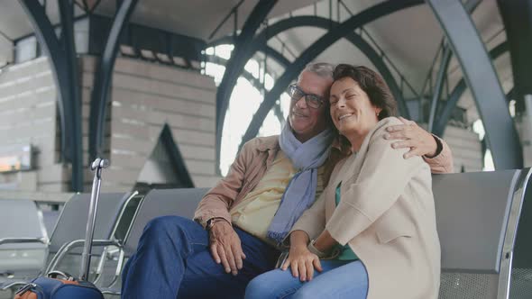 Mature Couple Hugging Sitting on Bench with Luggage Bags in Airport Waiting Area