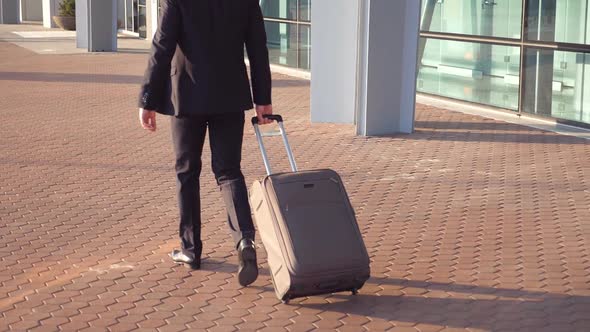 Rear View of Unrecognizable Young Businessman in a Suit Walking Near Terminal and Pulling Suitcase