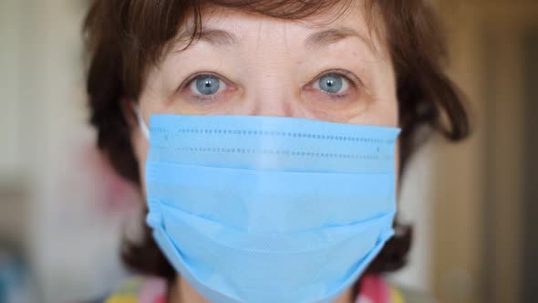 An aged woman in a protective medical mask is looking directly at the camera