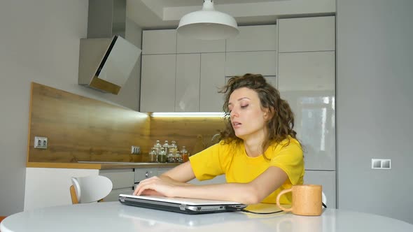 Tired Young Woman with Curly Hair and Yellow Shirt is Working From Home Using Her Laptop at the