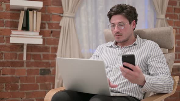 Middle Aged Man with Laptop using Smartphone on Sofa