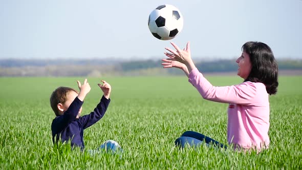 Happy Mom and Her Son Are Sitting on the Green Grass and Playing with the Ball, Throwing It Up 