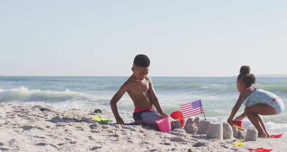 Smiling african american siblings building sandcastle with american flag on sunny beach