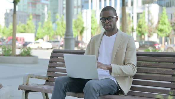 African Man with Laptop Smiling at Camera While Sitting Outdoor on Bench