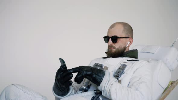 Astronaut Sits in a Chair with a Phone in His Hand and Listens to Music