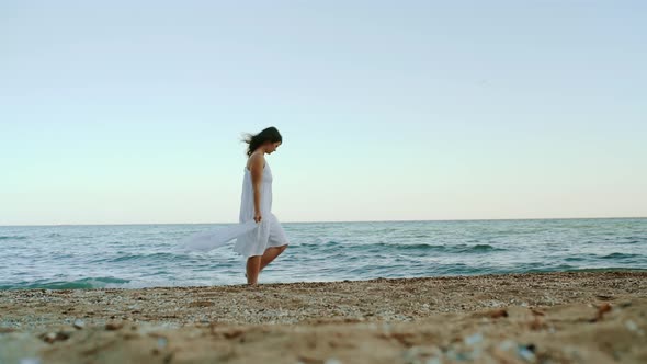 A Young Tourist Woman in a White Dress Walking Along the Coast of the Ocean