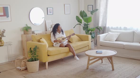 Cheerful Afro American Young Woman Playing the Ukulele Sitting on the Sofa