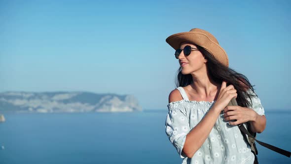 Smiling Travel Woman in Hat and Sunglasses Walking on Top of Mountain Over Sea Medium Shot