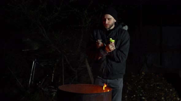 A Homeless Young Man Stands By the Fire and Eats an Apple. A Man Stands at Night Near a Barrel of