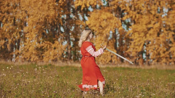 Woman in Red National Long Dress Standing on the Field and Fencing with a Sword