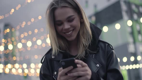 Young Girl Uses a Smartphone Sitting Outside in a Modern Residential Area Evening in the City Blurry