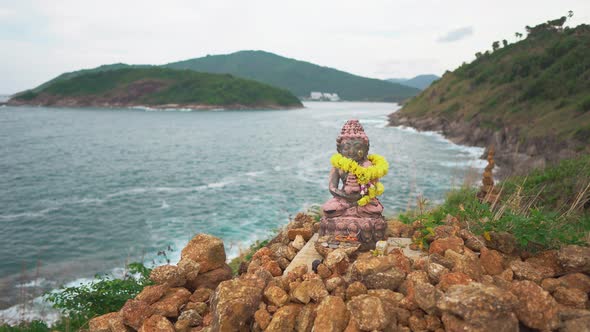 Little Statue of Sitting in A Lotus Position Deity on Top of A Cliff Against the Sea and Green Hills