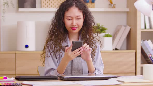 Happy Asian long curly hair woman scrolling on mobile phone at home office. Closeup joyful girl read